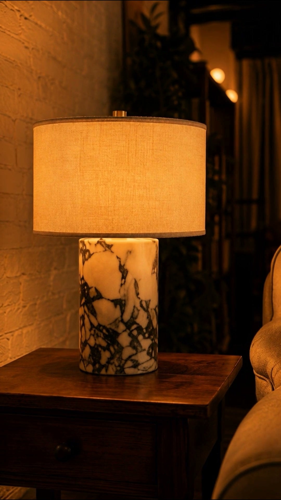 A marble table lamp lit on a side table against a brick wall